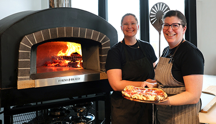 Two women taking pizza from large Italian ceramic oven